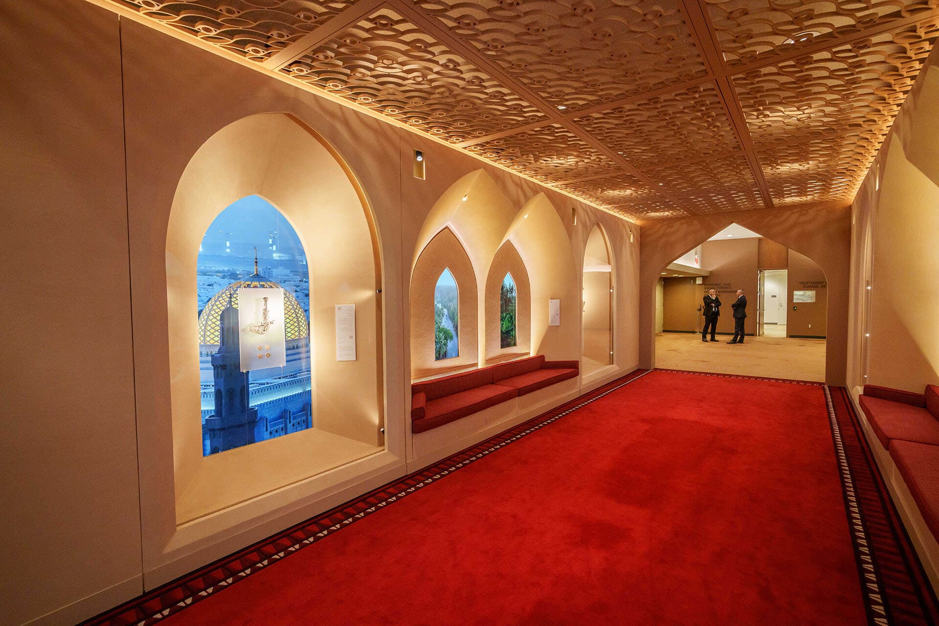 Interior del Corredor de Omán en la Sede de la ONU. La imagen muestra un pasillo con una vibrante alfombra roja y un techo de madera con intrincados patrones tallados. A la izquierda, una gran ventana arqueada muestra una imagen retroiluminada de la cúpula dorada de una mezquita. Más allá, se ven dos ventanas arqueadas más pequeñas con paisajes verdes. Un banco rojo tapizado se extiende a lo largo de la pared. Al fondo, dos figuras masculinas conversan cerca de una entrada.