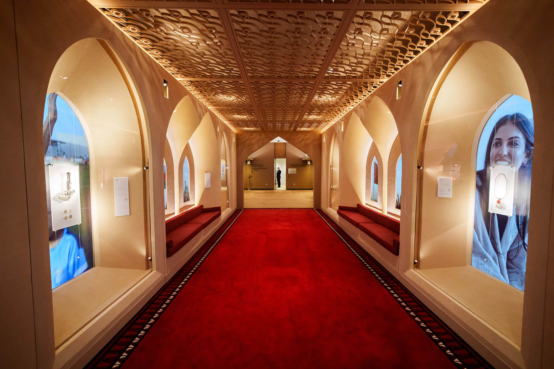 "Vista aérea del Corredor de Omán en la ONU. Muestra el detalle del techo de madera tallada con patrones geométricos iluminados durante una recepción. Hay una persona al fondo de pie, conversando.