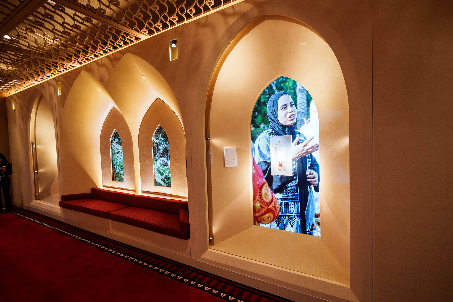 Vista lateral y detallada de las ventanas arqueadas del Display Sede Omán en la ONU. Muestra pantallas digitales con gráficos de alto contraste y un banco bajo tapizado en color burdeos.