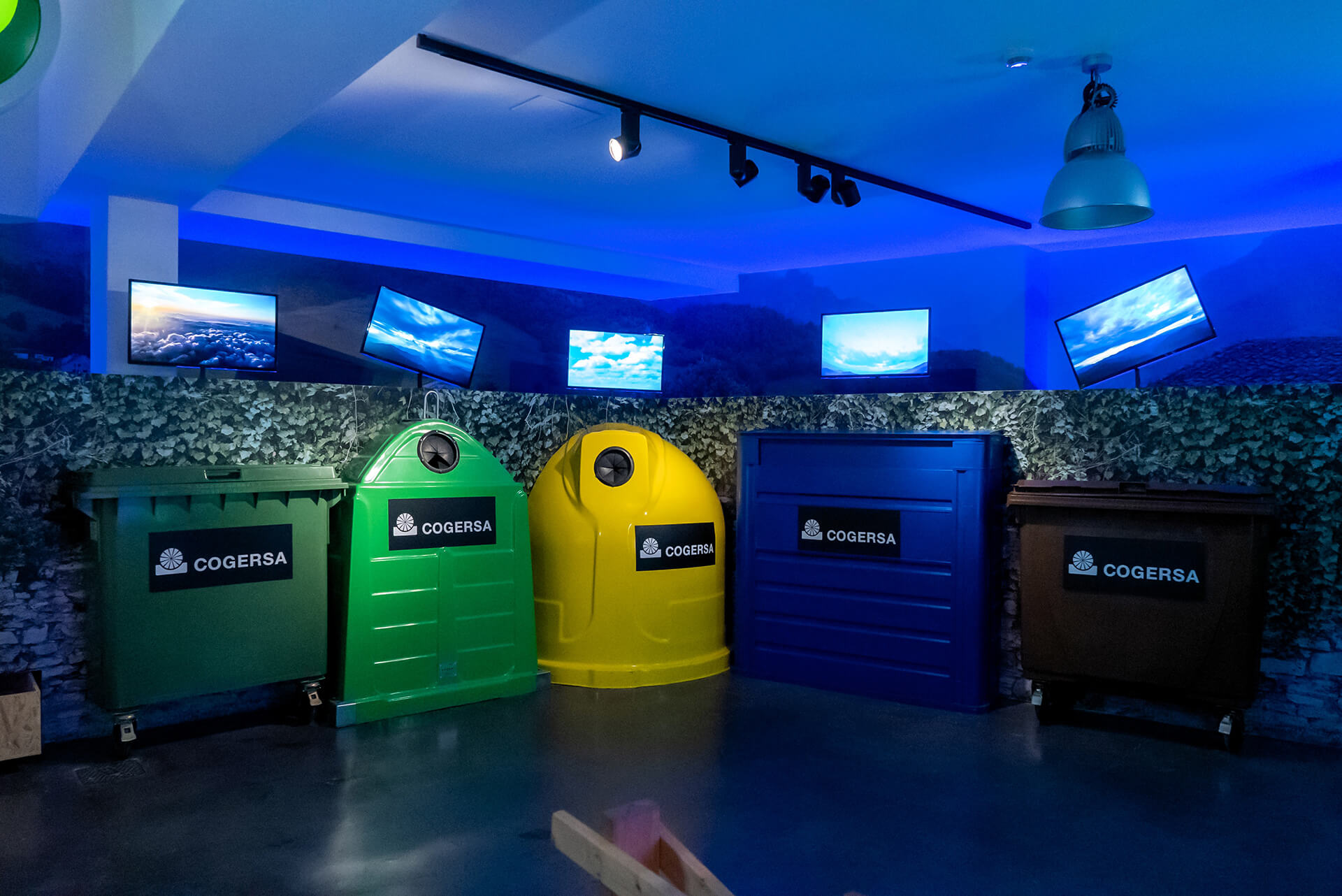 Sala con contenedores de reciclaje verde, amarillo y azul COGERSA bajo iluminación azul