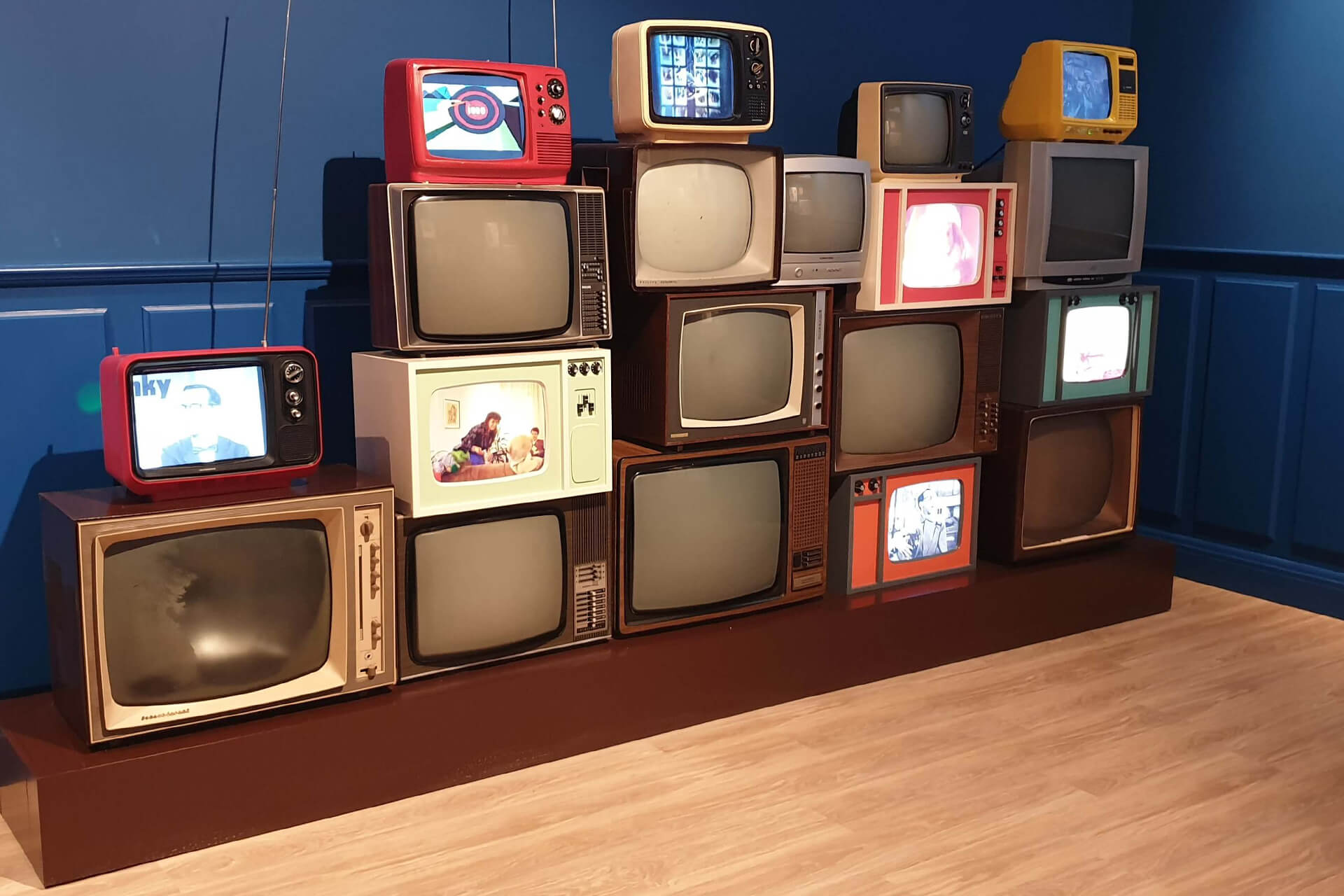 Conjunto de televisores vintage de distintas épocas sobre tarima de madera a medida, con pantallas encendidas retransmitiendo contenido sincronizado en The Chocolate Experience by 20|20, WOW Porto